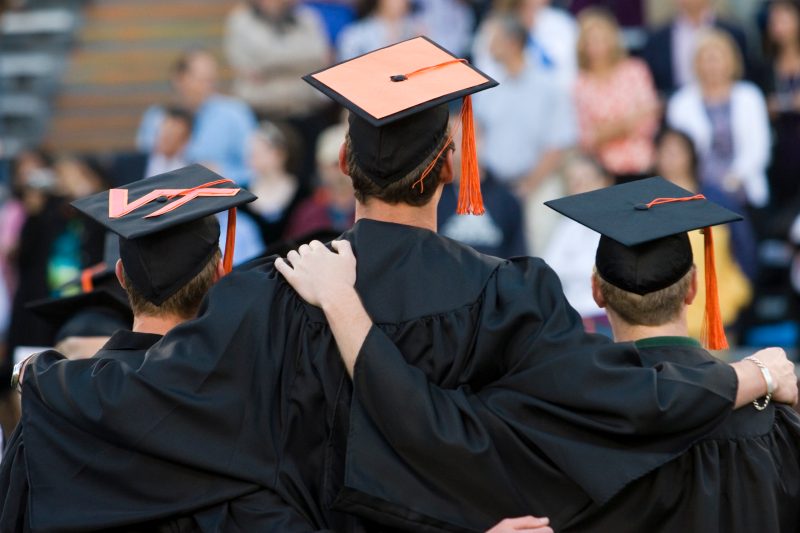 Friends stand at graduation with arms around one another's shoulders. They are wearing caps and gowns.